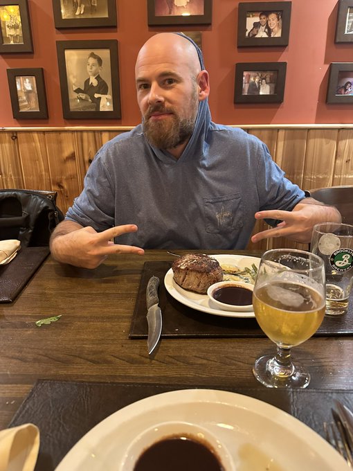 Casual photo of a bearded man wearing a blue hoodie pulled over his head, sitting at a restaurant table with a steak dinner, beer, and a knife in front of him.