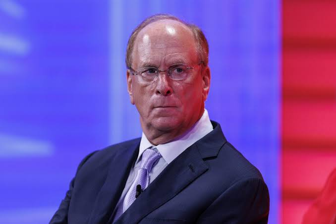 Larry Fink, CEO of BlackRock, is pictured in a dark suit with a purple tie against a red and blue backdrop, likely at a financial event. The image relates to the post about BlackRock's Bitcoin ETF selling $122.7M worth of BTC, highlighting the significance of institutional moves by the firm he leads.