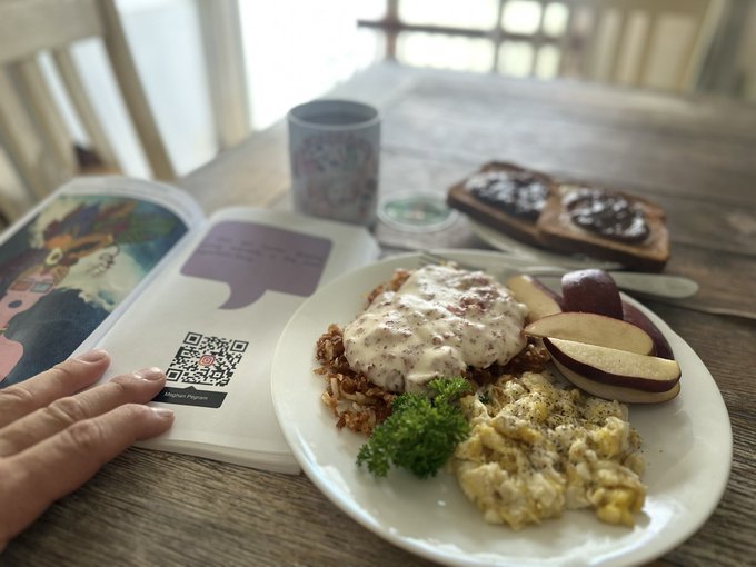 A first-person view of a hand holding open a children's book next to a hearty breakfast plate featuring scrambled eggs, hash browns topped with cream sauce, apple slices, and toast, with a mug on the wooden table.