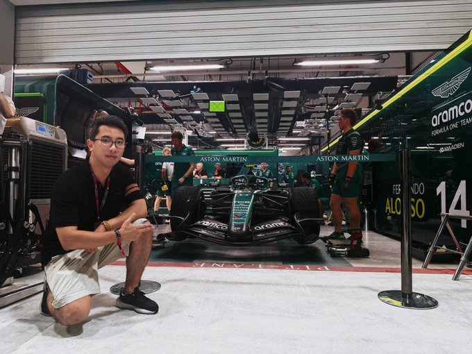 A man kneels in an Aston Martin Aramco Formula One Team garage in front of a race car, with team members working in the background.