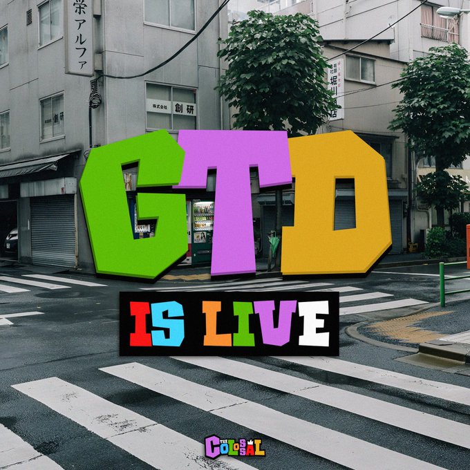 The image features a colorful sign that reads "GTD is now live." This message appears to be related to an event or promotion, possibly for a new album release by a popular artist. The sign is placed at the edge of a street with buildings in the background and a crosswalk nearby.