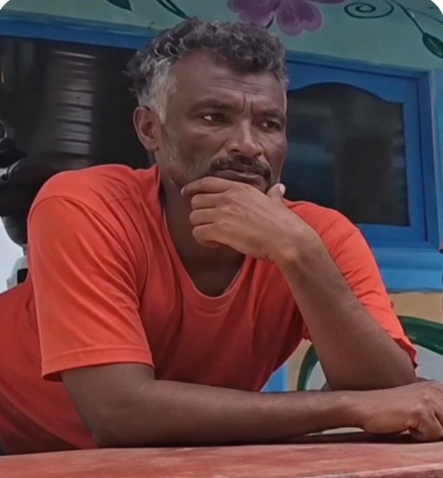 A contemplative middle-aged man with graying hair and a beard wearing an orange t-shirt, resting his chin on his hand with a pensive expression.