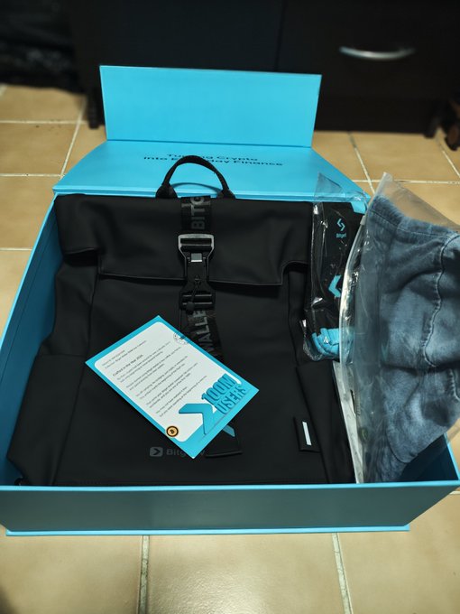 An open teal BitBasel gift box containing a black bag with branded straps, an information card, and other accessories on a tiled floor.
