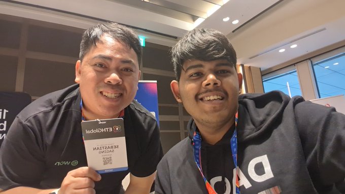 Two smiling men pose together at an event, one wearing a 'KALTZAB32 OFFICIAL' lanyard badge from 'LEADIQHTECH' and the other wearing a dark hoodie.
