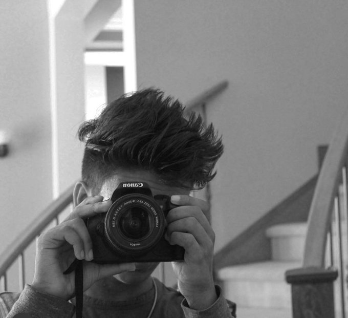 A black-and-white mirror selfie shows a person holding up a Canon EOS camera that obscures their face, their tousled hair prominent against a stairwell background. The camera's lens displays "CANON LENS EF 50mm 1:1.8 STM" and "Canon" branding, reflecting the "unfiltered" self-reflection theme of the crypto introduction post.