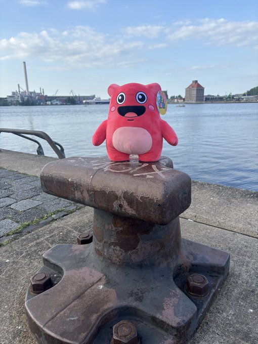 A pink plush toy with a smiling face sits atop a large rusty metal bollard on a waterfront promenade, with industrial buildings and water visible in the background.
