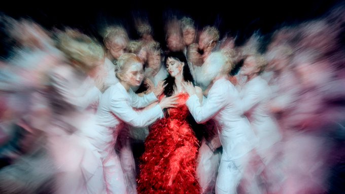 A central figure in a striking red gown is surrounded by blurred figures in white, creating a dreamlike, swirling composition that captures motion and energy—evoking themes of transformation and identity that resonate with the ethereal, collectible nature of digital art on Ethereum. The motion-blurred, otherworldly aesthetic suggests the intangible, decentralized spirit of blockchain-based artistic expression.