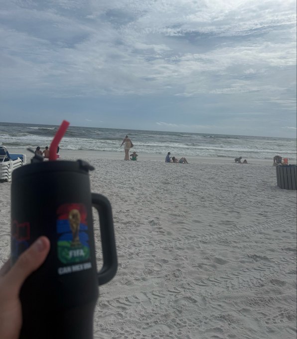 A hand holds a black tumbler cup featuring the 2022 FIFA World Cup Qatar logo while overlooking a sandy beach scene with people relaxing near the shore. The image juxtaposes a peaceful vacation moment with the humorous confession about suffering losses on memecoins, emphasizing the contrast between an outwardly relaxing getaway and hidden financial turmoil.

Visible text: "Maybe it's just me, but I think there is room for improvement here" and Chinese characters "自比张居正了？真是不白来中国..." along with "FIFA" and "Qatar 2022" branding on the cup.