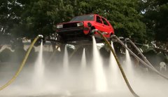 A red and black sports car sits elevated on ramps in a forested area, surrounded by streams of water spraying outward from underneath.