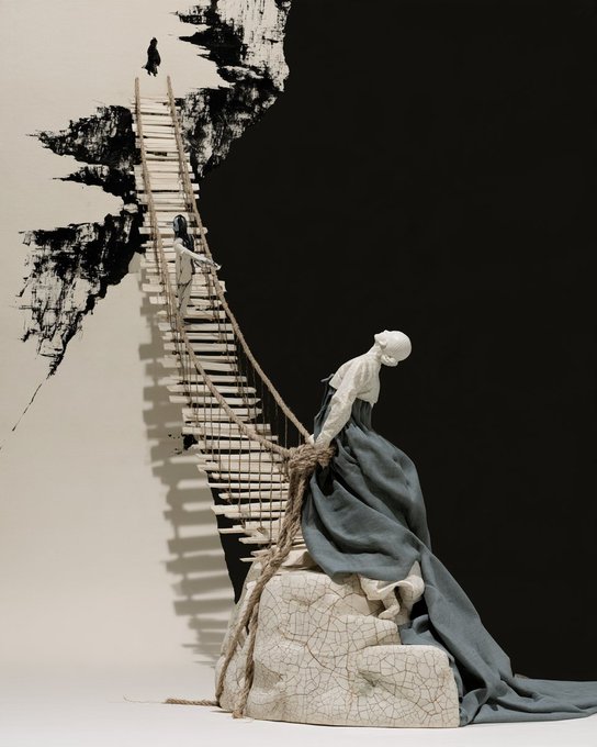 An artistic sculpture featuring a faceless figure in flowing dark and white robes sitting on a cracked block, with a wooden ladder/bridge extending upward toward a distant figure against a stark black and white background.