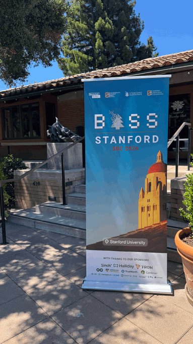 An outdoor banner for 'BOSS STANFORD GWC 2025' stands on a sidewalk near a building, displaying logos for sponsors including 1inch, Halliday, TRON, OKX, TrueNorth, and Candlen.