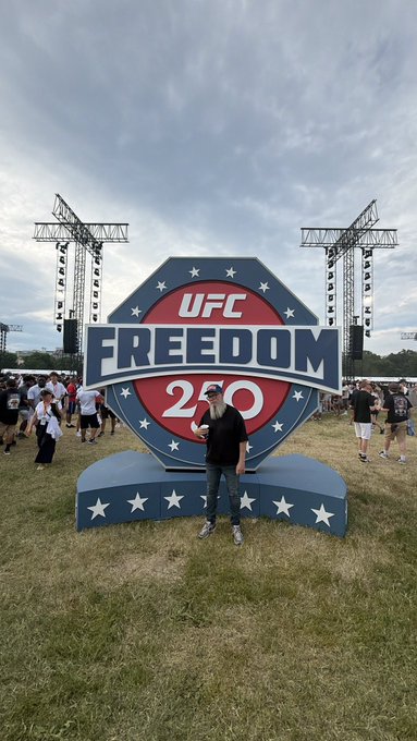 A man poses in front of a large UFC Freedom 250 sign at an outdoor event with stage rigging visible in the background.