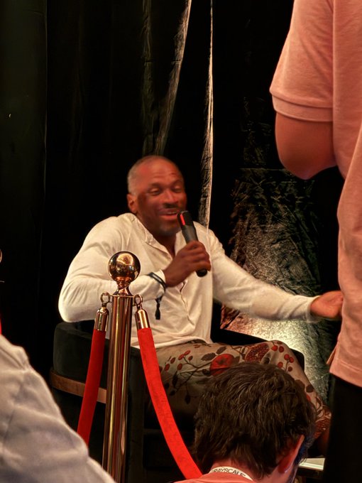 A man in a white button-down shirt sits in a chair holding a microphone, smiling during what looks like an interview or panel discussion, with a gold stanchion and red velvet rope in the foreground.