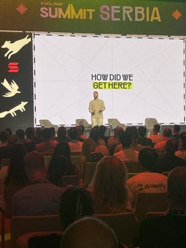 A speaker presents on stage at the 'SUMMIT SERBIA' event, with the slide behind him reading 'HOW DID WE GET HERE?' while attendees watch from the audience.