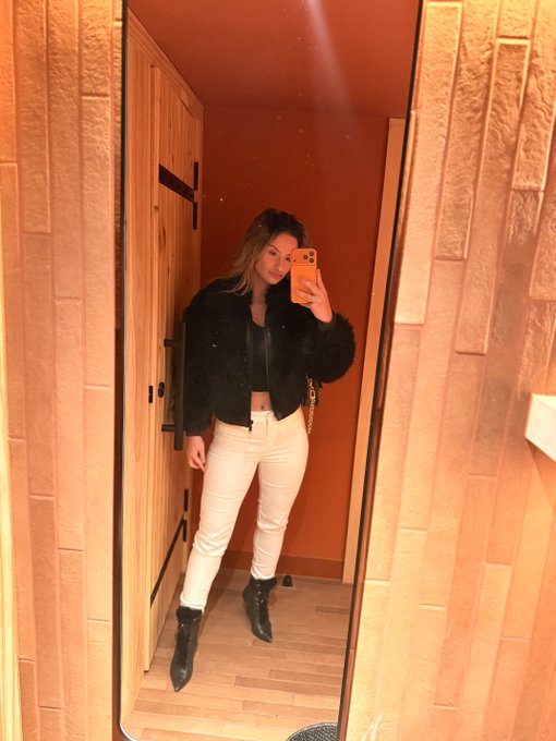 A mirror selfie of a woman in a black fuzzy jacket, white jeans, and black ankle boots, holding an orange phone in an orange-walled room.