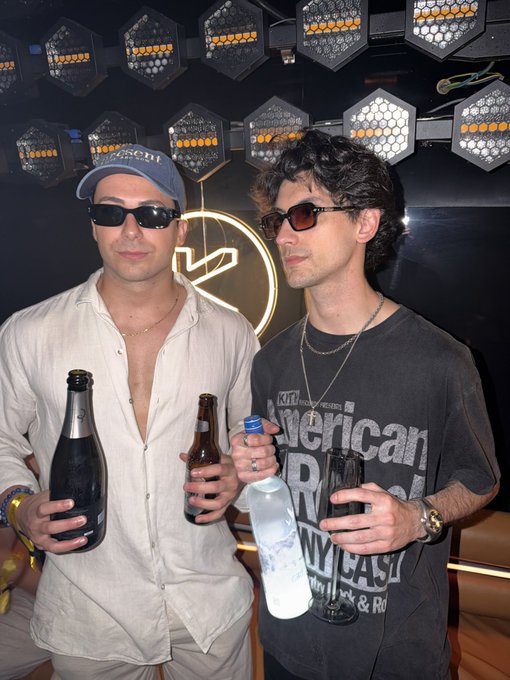 Two stylish men pose at what appears to be a nightclub or bar, both wearing dark sunglasses and holding drinks—one with a bottle of champagne and the other with a beer and water bottle. The man on the right sports a faded black "American" t-shirt and silver chains, fitting a "Solana Sensei" vibe of crypto-culture cool under the venue's hexagonal light fixtures.