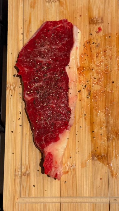 A raw, marbled steak seasoned with pepper rests on a wooden cutting board, glistening with juices.