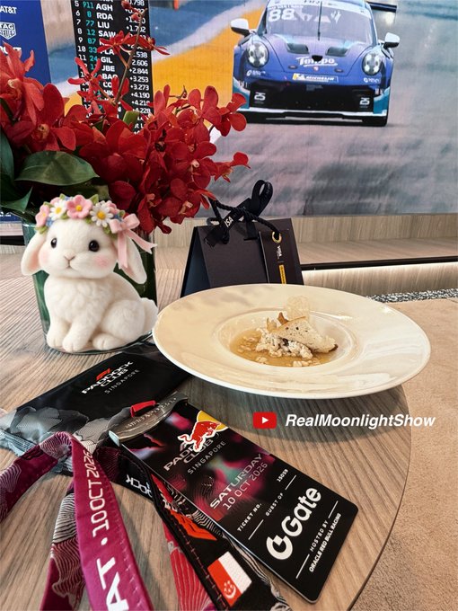 The image shows a Paddock Club VIP hospitality experience at the Singapore F1 race, featuring an elegant plated dish alongside Gate-branded merchandise (lanyard with "100CT202" and a "G Gate" sponsor card), a cute plush lamb figurine with flowers, and a TV in the background displaying a Porsche race car with the number 88, capturing the luxurious perks sponsored by Gate cryptocurrency exchange.