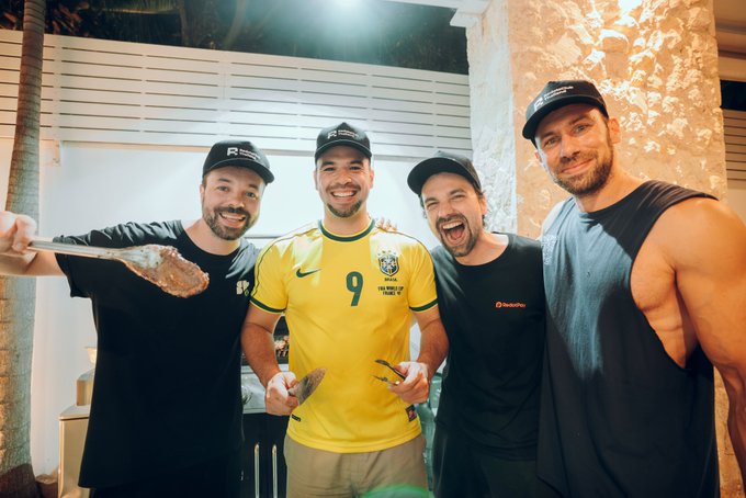 Four smiling men pose together at night; the man in the center wears a yellow Brazil national team jersey with the number 9, and one man holds a large spoonful of food.