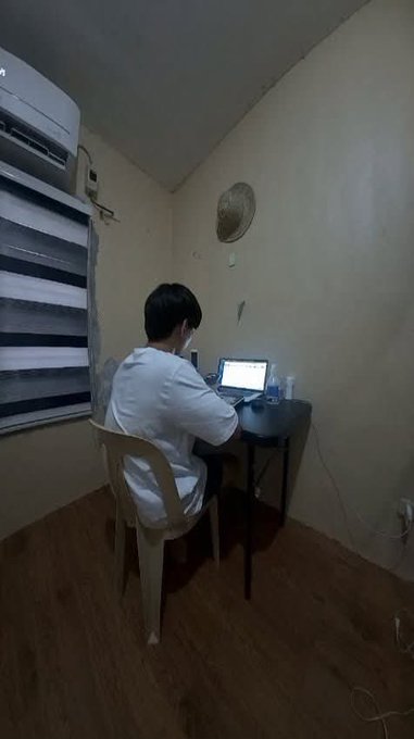 A person wearing a mask sits at a small desk working on a laptop in a dimly lit, narrow room with wooden floors and an air conditioner on the wall.