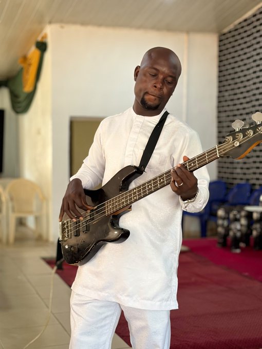 A man in a white traditional outfit plays a black bass guitar with his eyes closed, standing on a red carpet in what appears to be a hall.