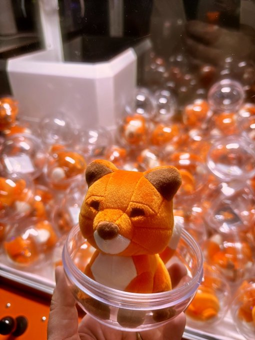 A hand proudly holds up a clear gachapon capsule containing an orange fox plushie—the MetaMask mascot—which appears to have been won from one of the capsule machines visible in the background, marking it as one of the very first MetaMask plushies ever produced.