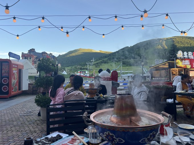 An outdoor dining scene at a hot pot restaurant, where people are enjoying steaming copper hot pots under string lights with mountains in the background, capturing the lively atmosphere that contrasts with the poster's frustration over declining crypto investments despite an otherwise perfect day.