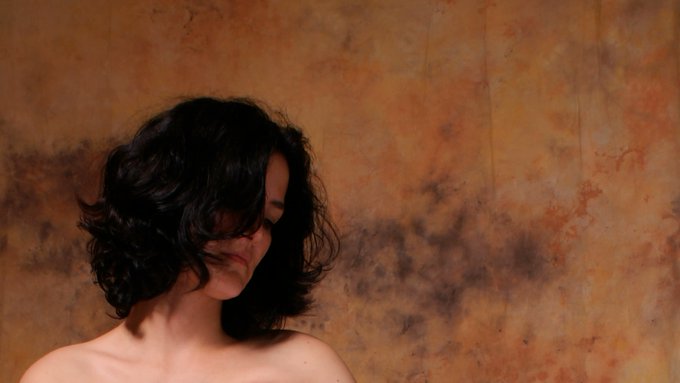 A bare-shouldered woman with dark, tousled hair partially obscures her face, set against a richly textured, rusted orange-brown wall that echoes the warm, weathered tones of the post's theme of artistic curation. The composition reflects the contemplative, minimalist aesthetic highlighted in the featured work "Cut Wires" by @m_blehaut.