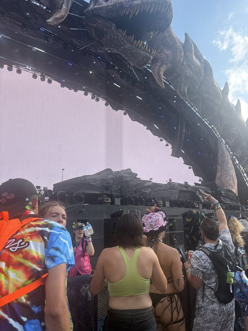 A festival crowd scene with attendees in colorful rave attire watching a large stage featuring a massive dinosaur head sculpture, with a giant blank screen and stage set visible.