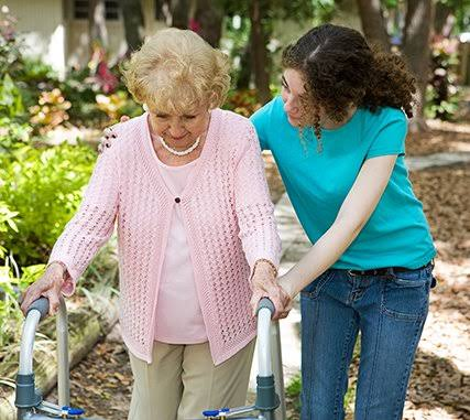 An elderly woman uses a walker with assistance from a younger woman in a teal shirt in an outdoor garden setting.