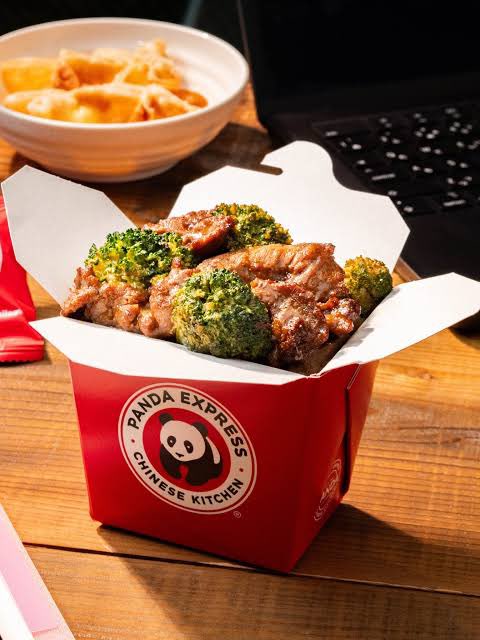 A red Panda Express Chinese Kitchen takeout box filled with beef and broccoli, served alongside a bowl of orange chicken next to a laptop keyboard.