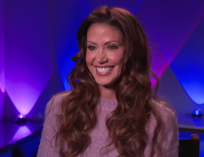 A smiling woman with long, wavy brown hair wearing a light purple sweater appears on a television set with purple and blue lighting.