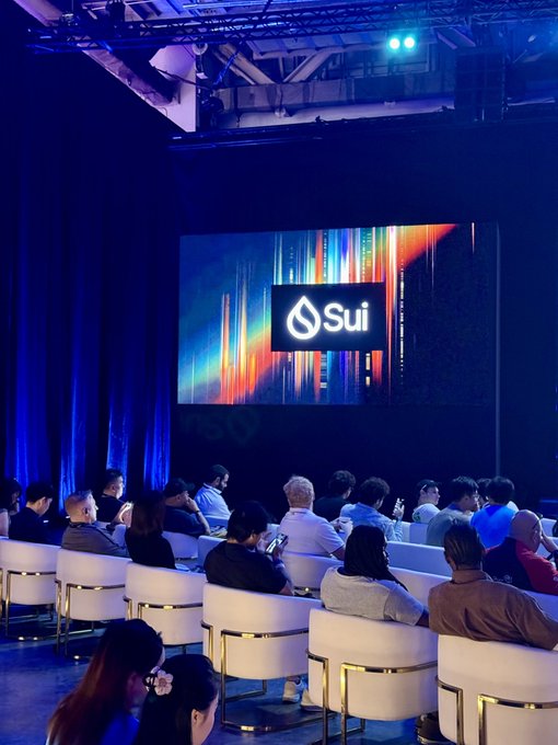 Attendees gather in a dimly lit auditorium at Sui Basecamp, with a large screen prominently displaying the "Sui" logo and colorful graphics, capturing the tech event's professional atmosphere. The seated audience, some holding phones, reflects the engaged community celebrated in the post.