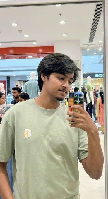 A young man takes a mirror selfie in a shopping mall while wearing a light green t-shirt and holding a phone with a colorful case, and the caption "Me: My Fav Wallet:" humorously compares the empty look of his pants pockets to his sleek phone case.