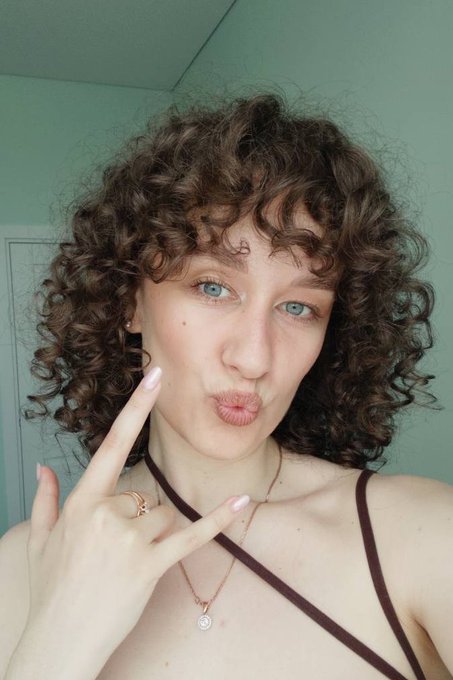 A young woman with curly brown hair poses playfully, making a peace sign and puckering her lips while wearing a brown top with thin straps and layered necklaces.