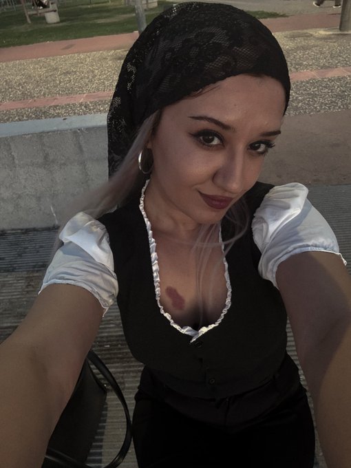 A selfie of a young woman wearing a black lace headscarf and a black vest over a white blouse with puffed sleeves, sitting on a bench outdoors.