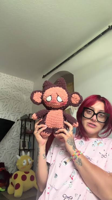 A woman with pink hair and glasses holding up a handmade crocheted monkey plushie, with a yellow giraffe plush visible in the background.