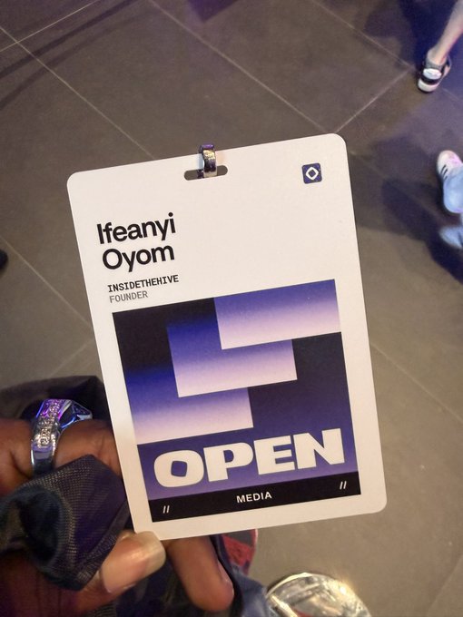 A close-up photo of an event badge being held by hand, displaying the name 'Ifeanyi Oyom', the title 'INSIDETHEHIVE FOUNDER', and the words 'OPEN MEDIA' over a purple gradient background.