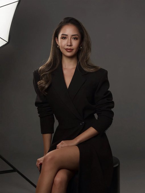 A woman with long wavy brown hair sits in a professional studio portrait, wearing a black blazer dress and pearl earrings, with a softbox light visible to the side against a dark backdrop. The polished, conference-ready look complements the post's announcement of her attending Token2049 Singapore to present insights from an upcoming RWA report with Dune.