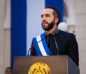 A bearded man wearing a blue and white striped presidential sash speaks at a podium bearing a gold seal.