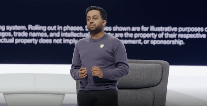 A man in a purple sweater stands on a stage giving a presentation, with partially visible text behind him including 'system. Rolling out in phases' and 'illustrative purposes'.