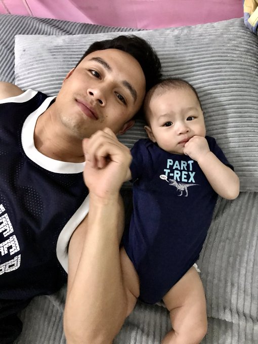 A man and a baby lying side by side on a striped cushion; the baby's blue onesie reads 'PART T-REX'.
