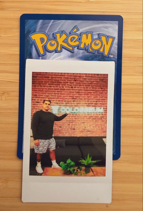 A Pokémon trading card placed on a wooden surface, featuring a photo of a man in a black sweater and shorts standing in front of a 'COLOSSEUM' sign against a brick wall.