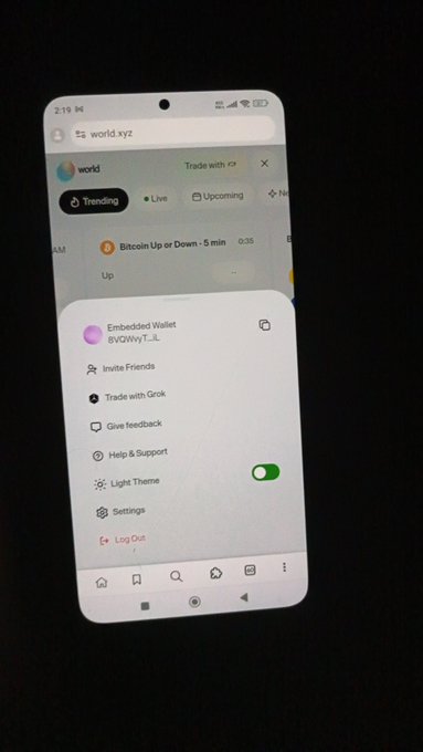 A smartphone displays the world.xyz app with an open menu showing options like "Embedded Wallet" (BVGYWY...il), Invite Friends, Trade with Grok, Give feedback, Help & Support, Light Theme, Settings, and Log Out, against a backdrop of a "Bitcoin Up or Down · 5 min" prediction market. The image directly supports the post by showcasing the active platform being highlighted as @world_xyz prepares to launch its reward feature.