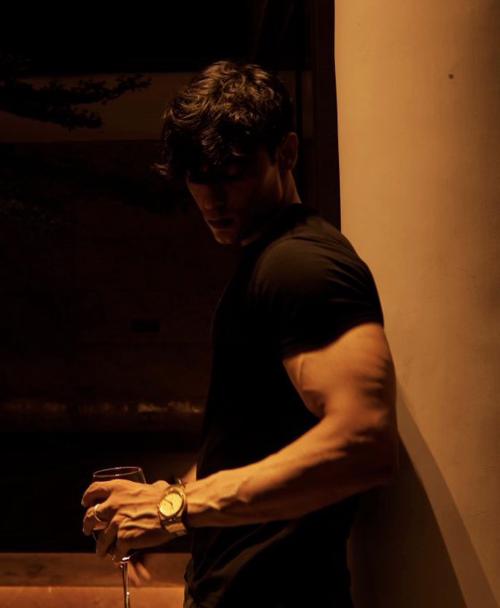 A man with dark hair wearing a black t-shirt stands against a wall in dim lighting, holding a wine glass and wearing a gold watch.