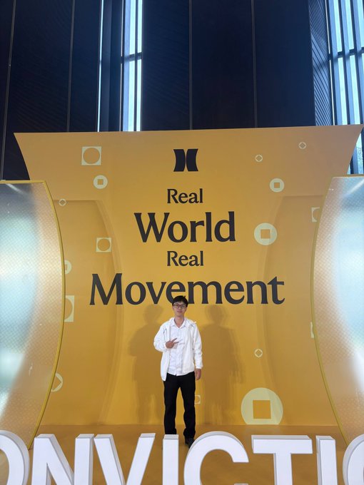A man in a white shirt stands in front of a yellow backdrop displaying 'Real World Real Movement' and 'INVICTUS' branding.