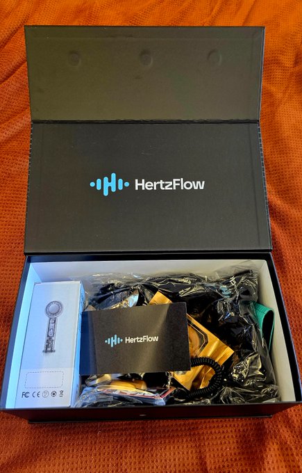 An open black box displaying the brand name 'HertzFlow' on the inner lid, containing a small white electronic device and gold packaging materials.