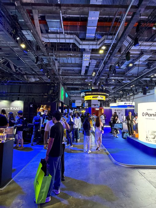 A crowded indoor exhibition hall features booths including 'AST' and 'OPanic', with attendees walking on blue carpeted flooring.