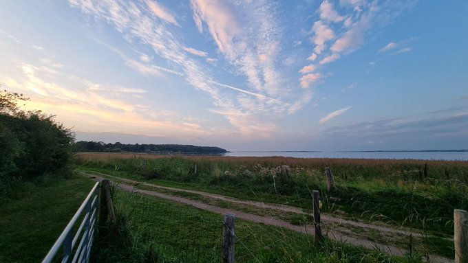 The image shows a beautiful view of a lake surrounded by grassy fields, with a fence running along its edge. The sky above has a mix of blue and white clouds, creating a serene atmosphere. In the foreground, there are two wooden posts standing next to each other, possibly part of a fence or gate. The scene is captured from behind these posts, giving an interesting perspective on the landscape.
