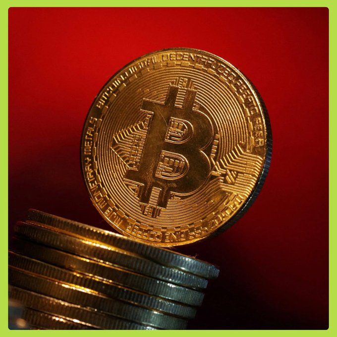 Close-up photograph of a gold Bitcoin coin standing upright on a stack of silver coins against a red background, framed by a yellow border.